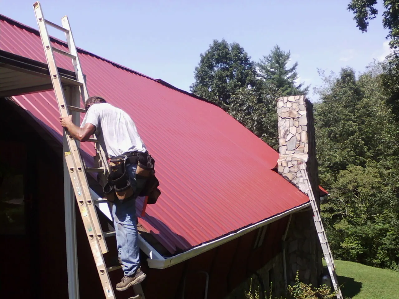 Roofer on a ladder beside a red metal roof and stone chimney — Soffit & Fascia Repair by Roofing Brothers in Woodhaven